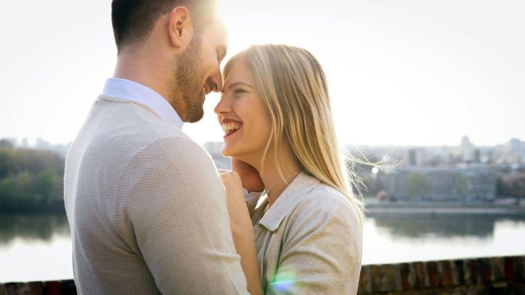 A man and a woman stand close, smiling at each other with sunlight in the background.