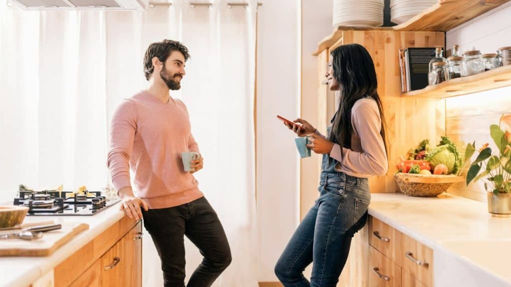 A man and a woman, both holding mugs, talk and lean against kitchen counters.