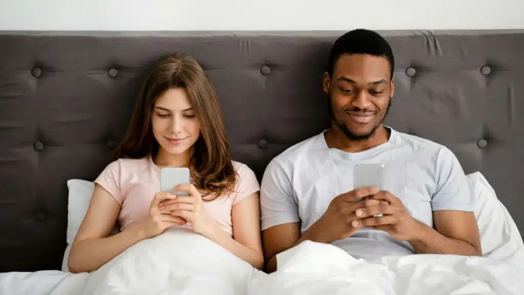 A woman and a man lie in bed, both looking at their phones.