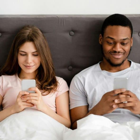 A woman and a man lie in bed, both looking at their phones.