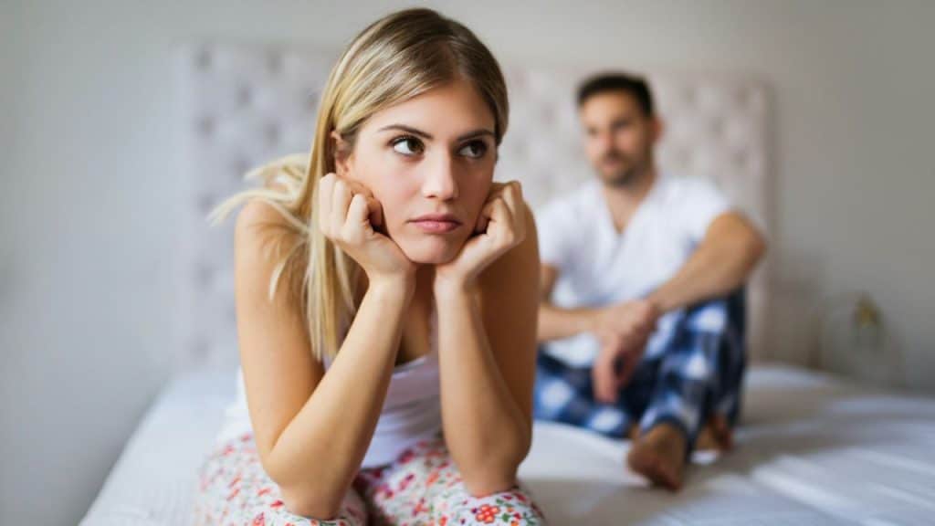 A woman with blonde hair sits on a bed, looking upset, with a man blurred in the background.