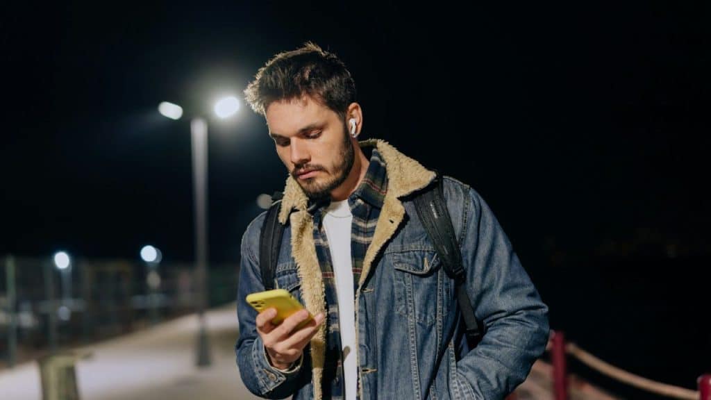 A bearded man in a denim jacket and white earbuds looks at his phone at night.
