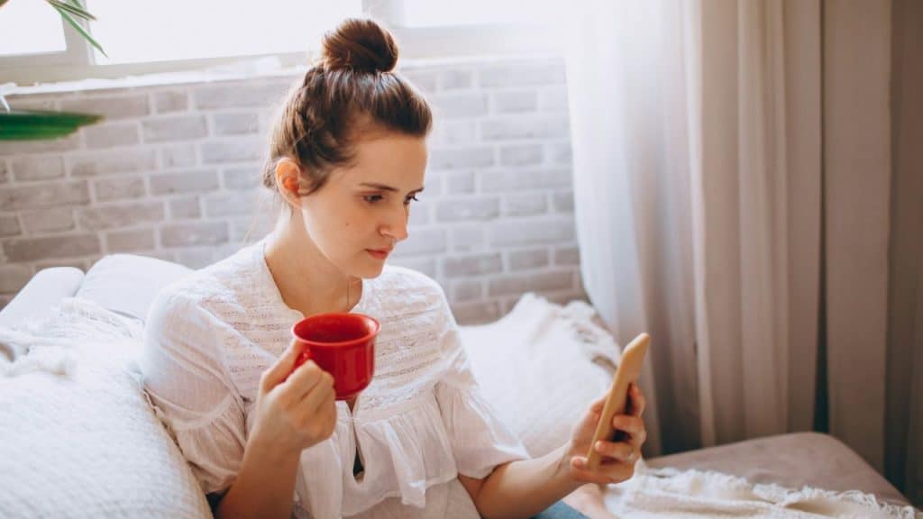 A woman with her hair in a bun holds a red mug and looks at her phone.