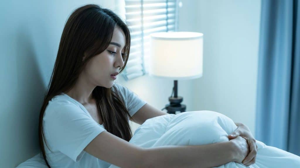 A young woman with long dark hair sits in bed, holding a white blanket, looking sad.