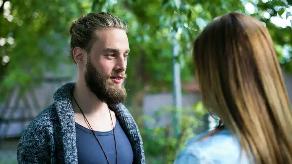 A man with a beard and hair tied back talks to a woman with long hair, seen from behind.