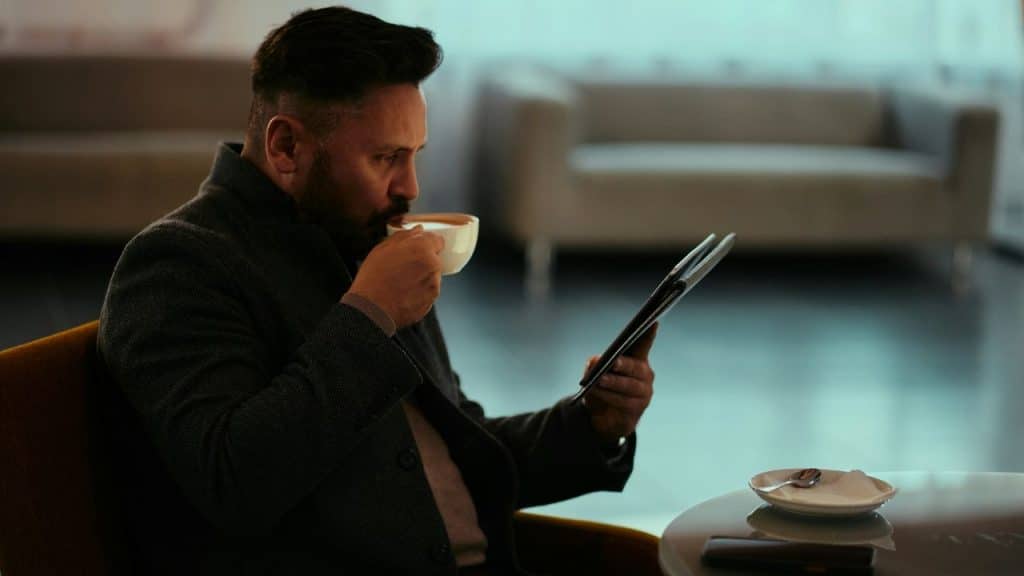 A man with a beard sips from a cup while reading a tablet in a modern setting.