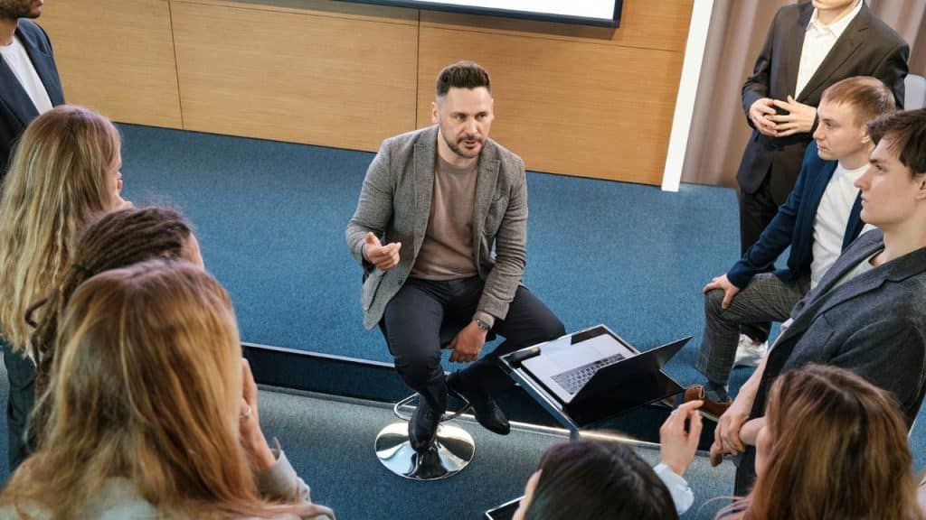 A man sits on a stool, talking to a group of people, with a laptop in front of him.