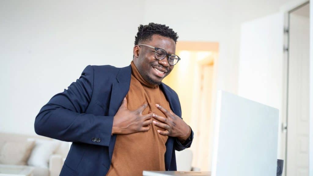 A happy man with glasses holds his chest while looking at a laptop, smiling.