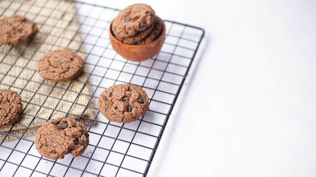 A wire cooling rack topped with chocolate chip cookies and a small wooden bowl filled with extra cookies.