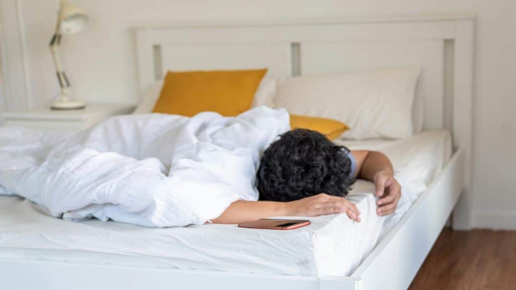A person face down under a white duvet, smartphone on the bed’s edge.
