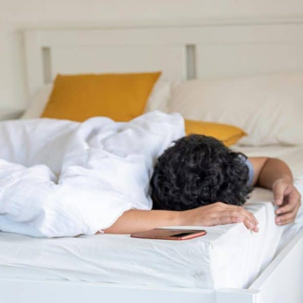 A person face down under a white duvet, smartphone on the bed’s edge.