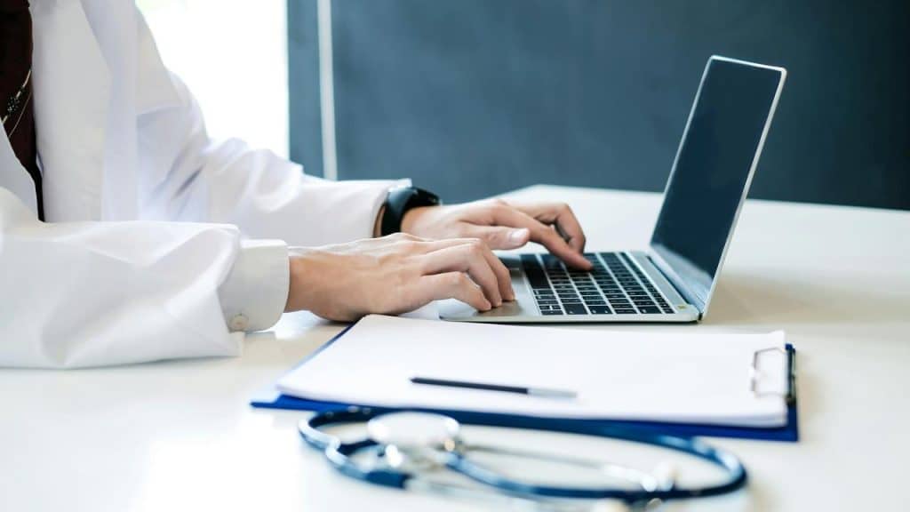 A doctor’s hands typing on a laptop beside a stethoscope and a clipboard.