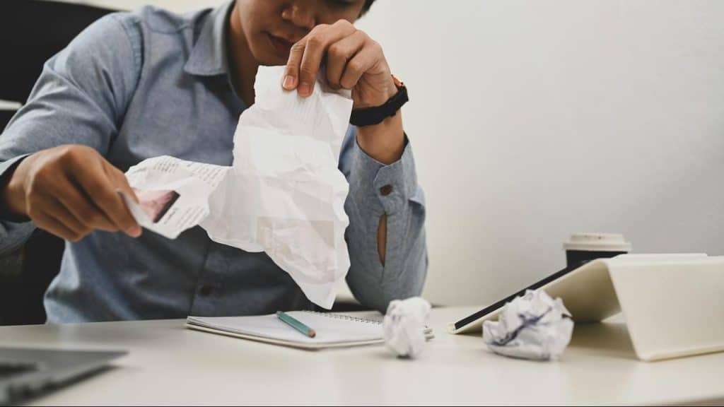 A person tearing up a sheet of paper at a desk with crumpled scraps nearby.