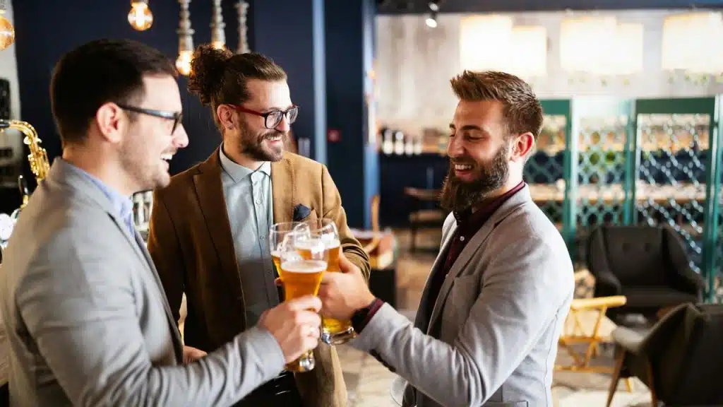 A group of three well-dressed men clinking glasses of beer and smiling in a stylish, modern bar.