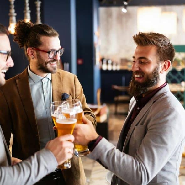 A group of three well-dressed men clinking glasses of beer and smiling in a stylish, modern bar.