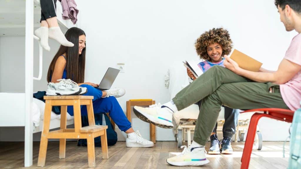 A group of three teens reading, working on a laptop, and chatting in a bright dormitory room.