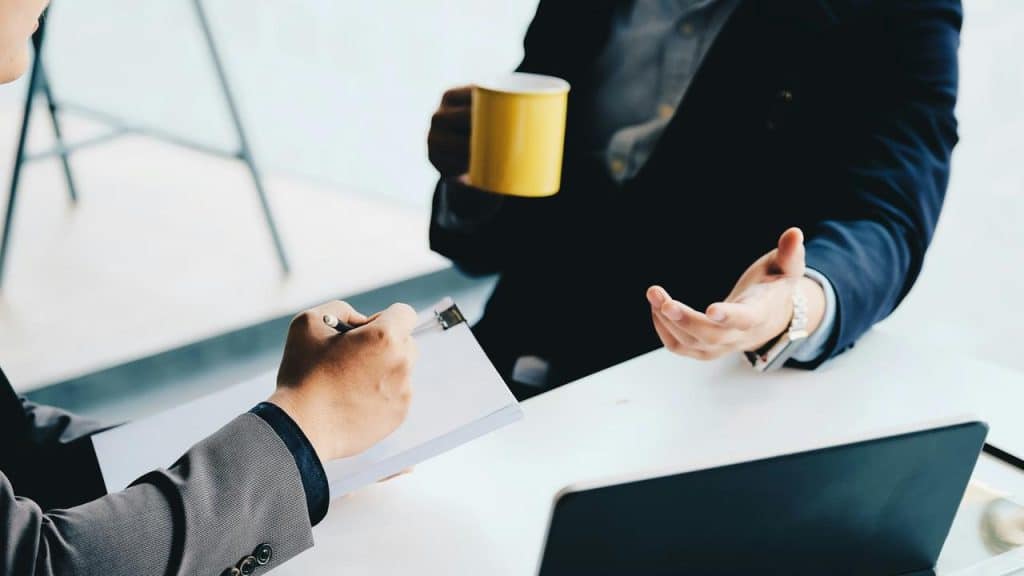 A person holding a yellow mug and gesturing while another takes notes on a clipboard at a desk.