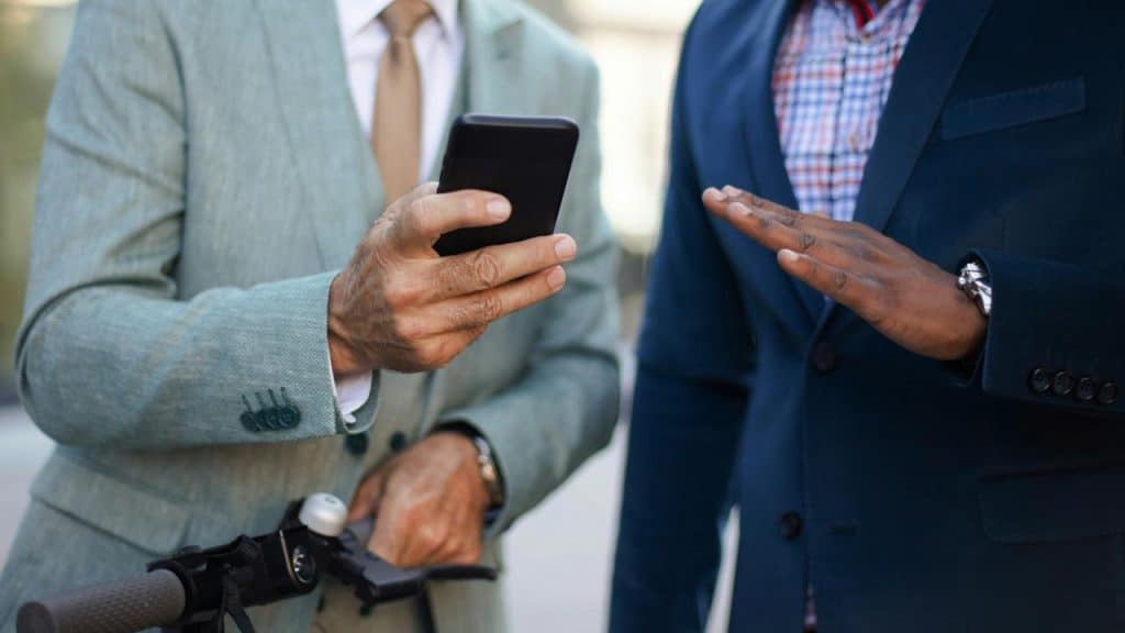 A man in a suit on a scooter showing his smartphone to another suited man.