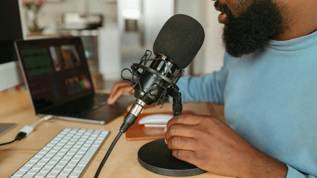 A bearded person adjusting a black studio microphone on a desk next to a laptop and keyboard.