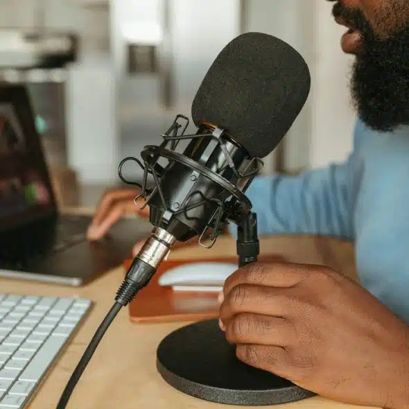 A bearded person adjusting a black studio microphone on a desk next to a laptop and keyboard.