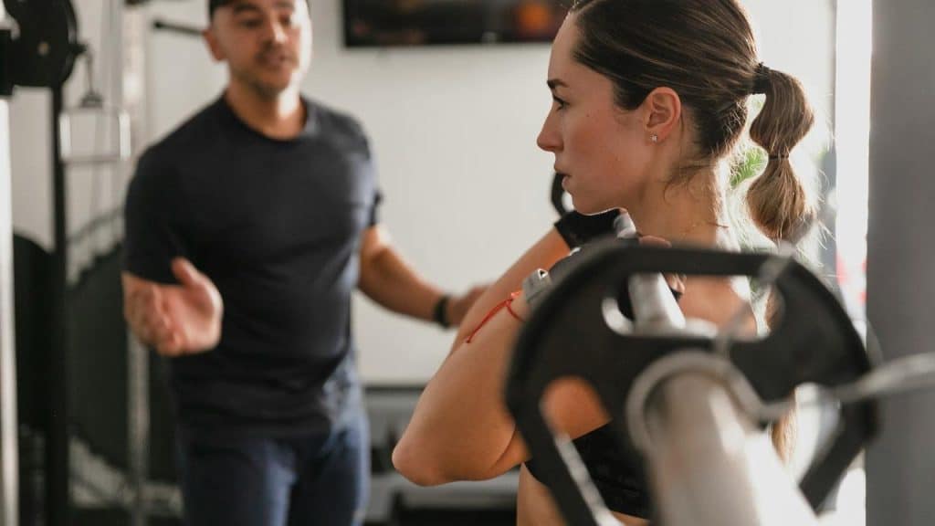 A woman lifting a barbell while a trainer watches and gives instructions.