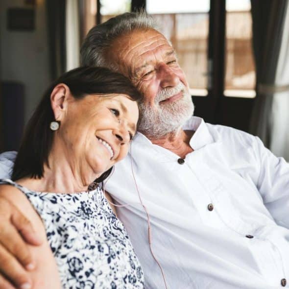 A smiling elderly couple embraces on a couch, looking content.