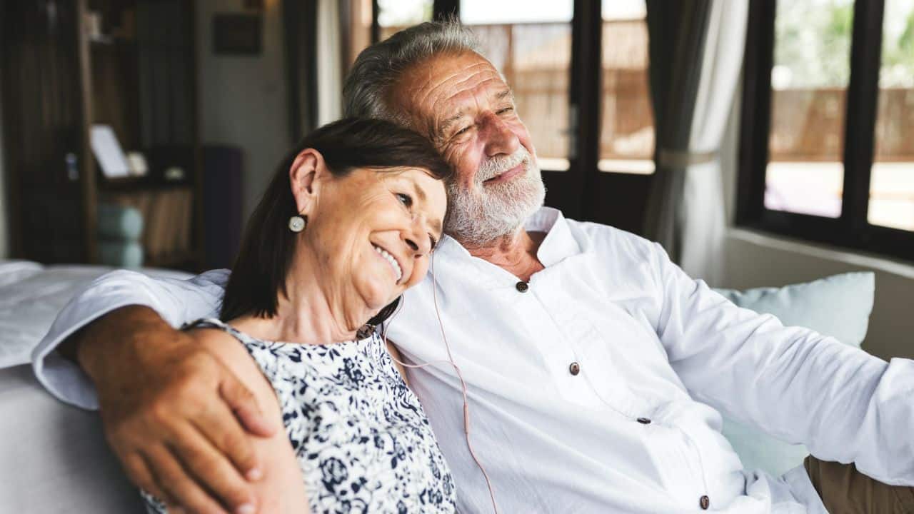 A smiling elderly couple embraces on a couch, looking content.