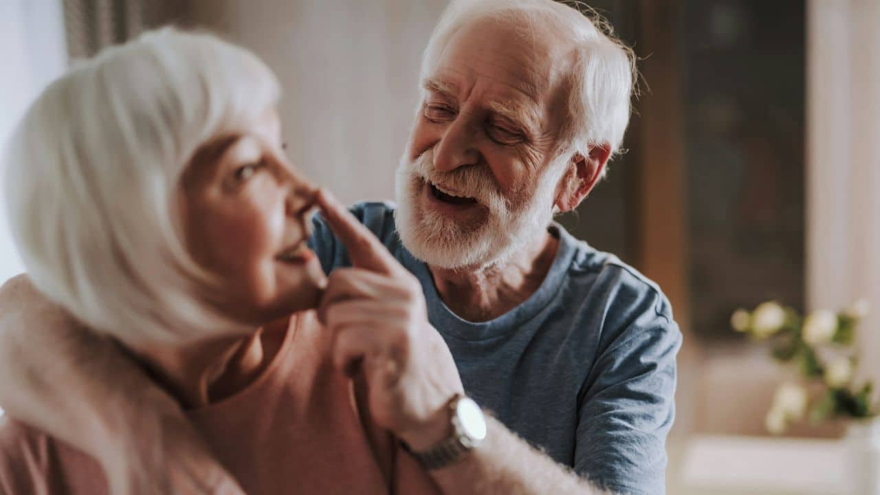 A happy elderly man with a white beard touches a smiling elderly woman's nose.