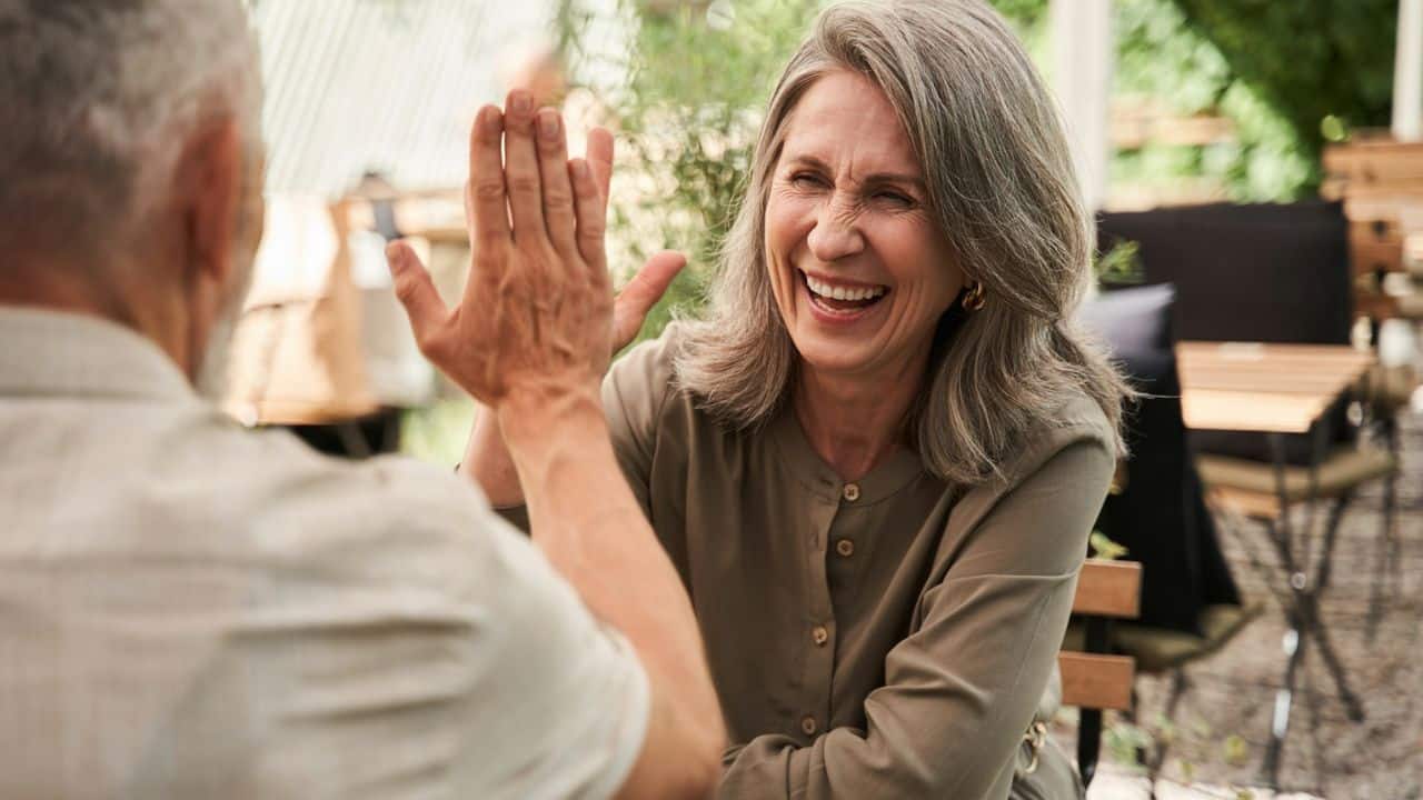 A laughing elderly woman high-fives a man at an outdoor cafe.