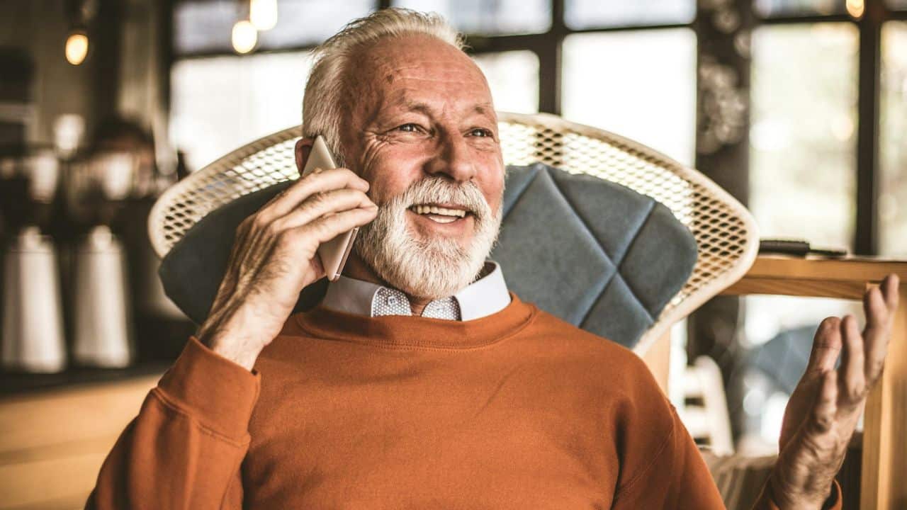 A smiling elderly man with a white beard talks on a cell phone, gesturing with his left hand.