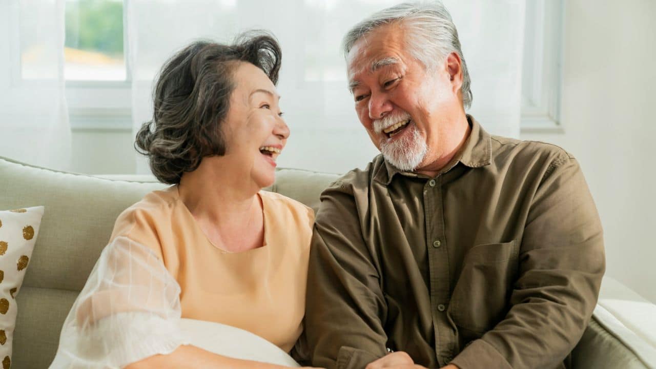 An elderly Asian couple sits on a couch, laughing and looking at each other.