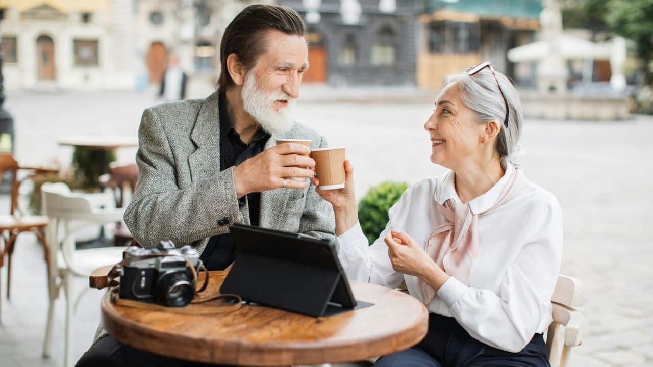An elderly couple smiles and clinks coffee cups at an outdoor cafe table with a camera and tablet.