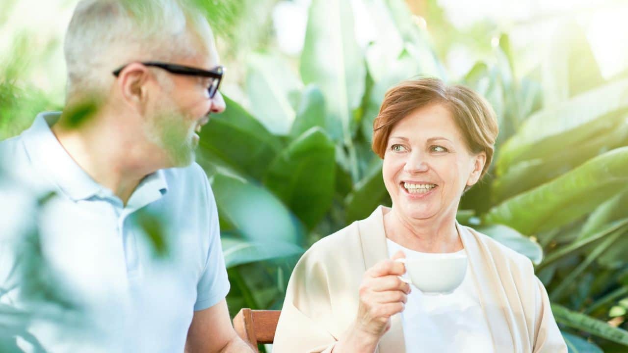 A smiling elderly woman holds a white cup and looks at a man with glasses.