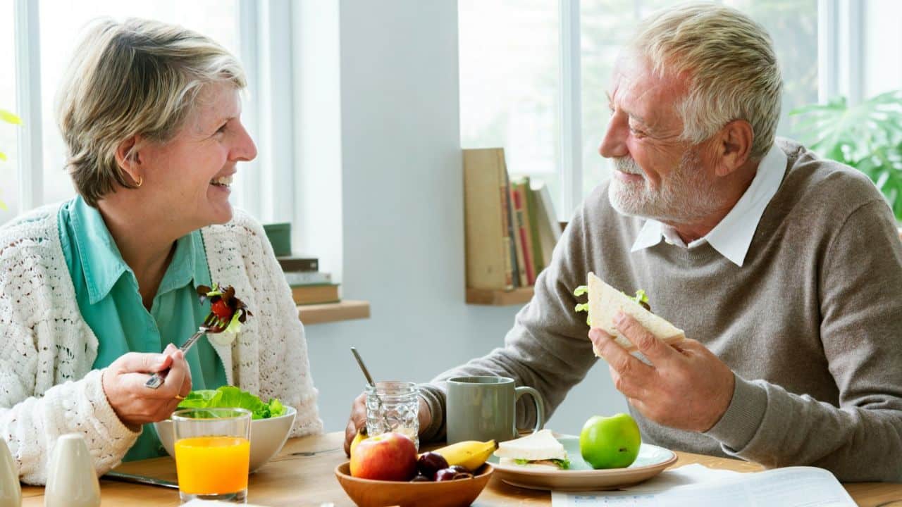 A smiling elderly couple enjoys a meal at a table with fruit and drinks.
