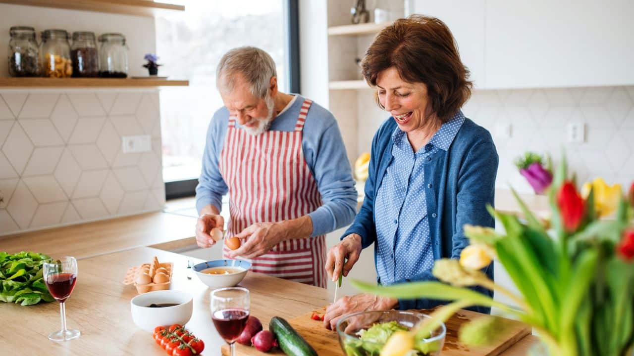 A happy elderly couple cooks together in a bright kitchen, with the woman chopping vegetables and the man cracking eggs.