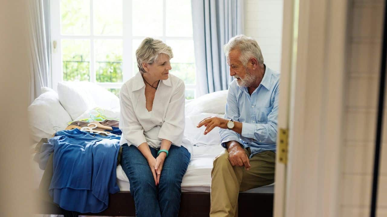 An elderly couple sits on a bed, talking, with clothes on the bed between them.