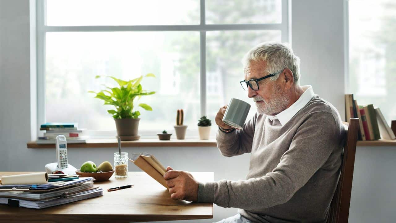 An elderly man with glasses and a beard reads a book, holding a mug.