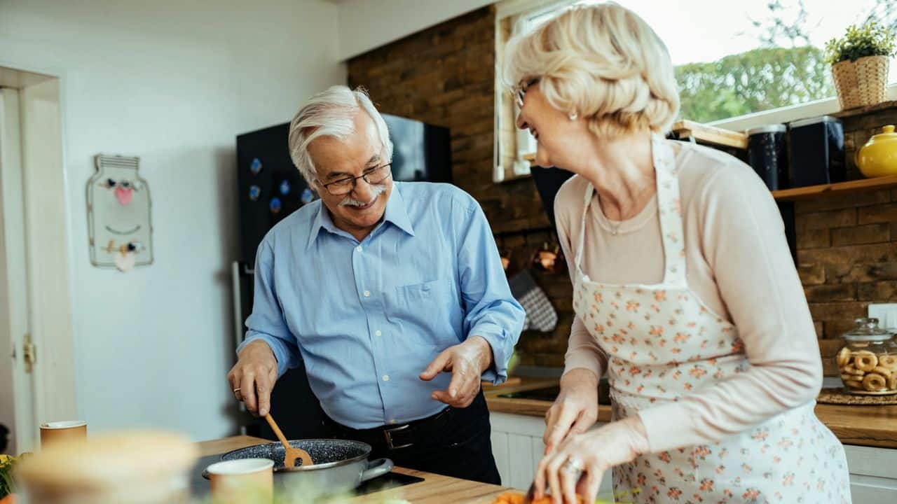 An elderly couple cooks in a kitchen, with the man stirring a pot and the woman chopping.