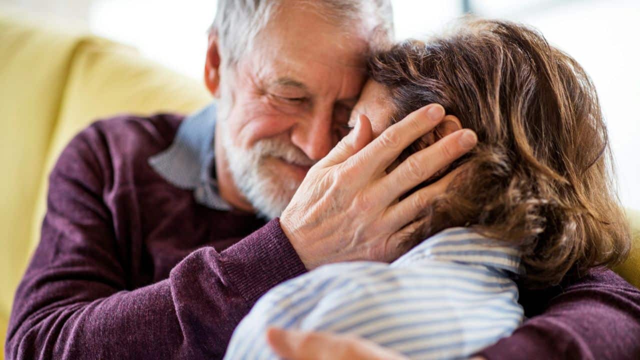 A man with a beard embraces a woman, tenderly touching her face, both smiling.