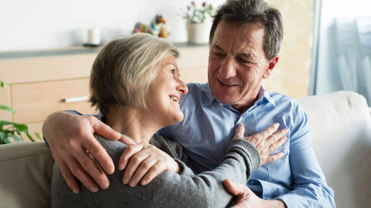 A smiling elderly couple embraces on a couch, looking at each other warmly.