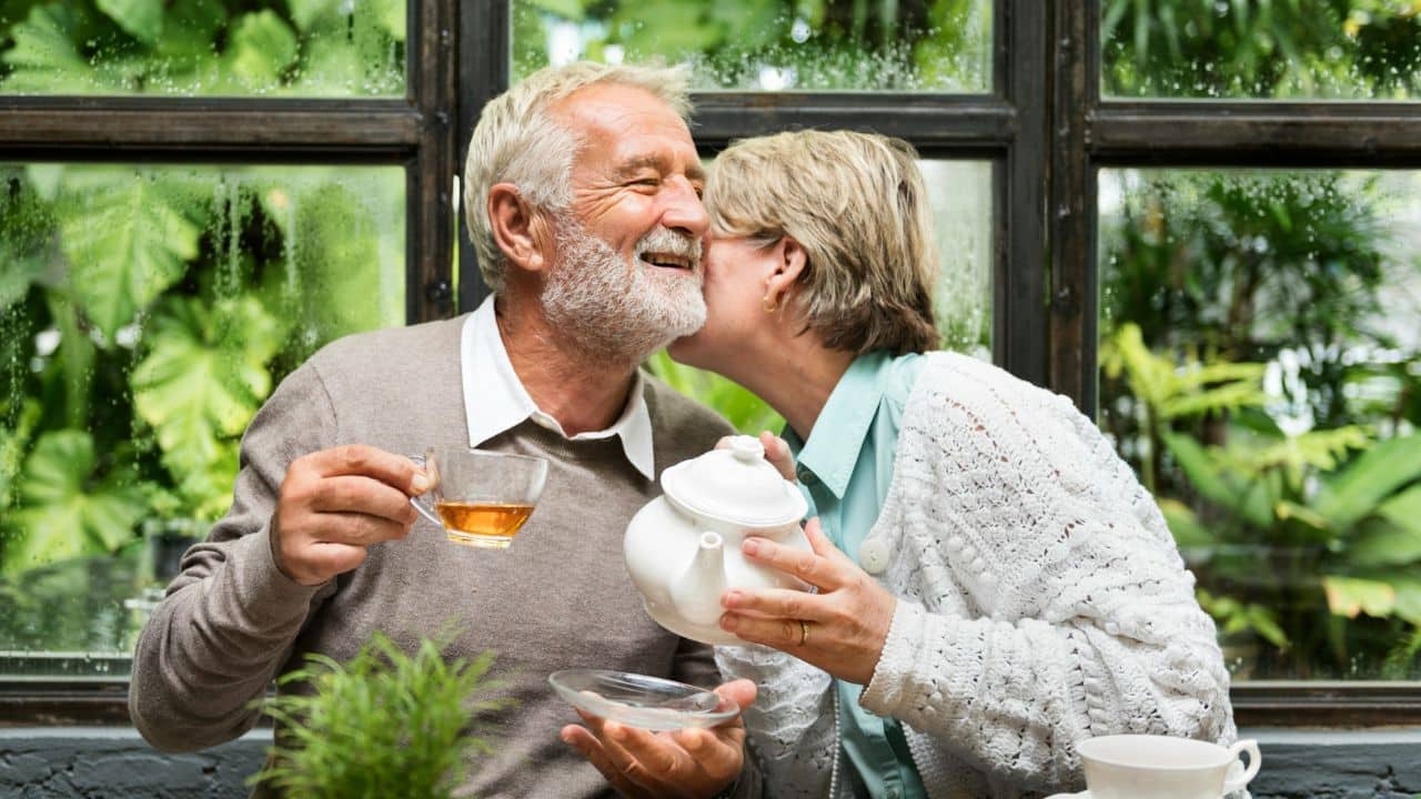 An older woman kisses a smiling older man's cheek while he holds a teacup.