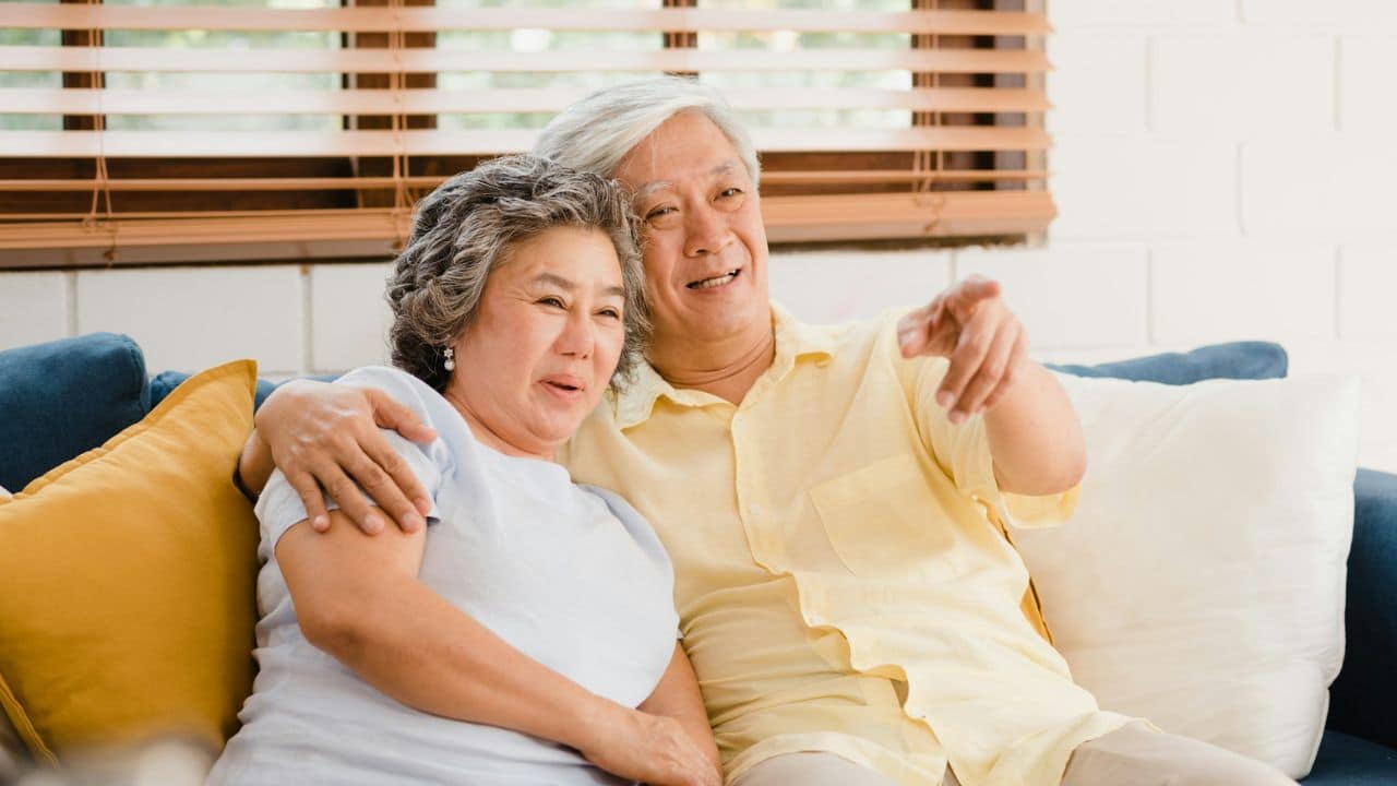 A smiling senior Asian couple sits on a couch, with the man pointing outwards.