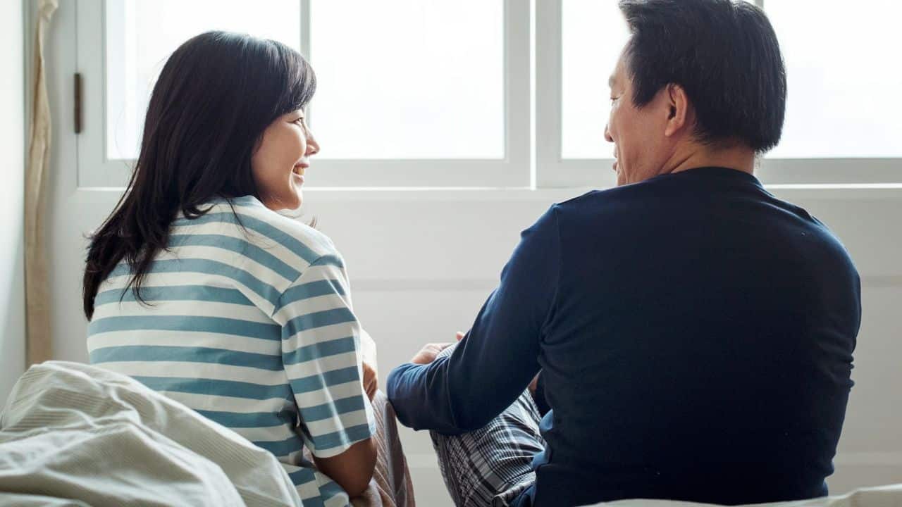 A smiling woman and a man sit facing each other on a bed, talking.