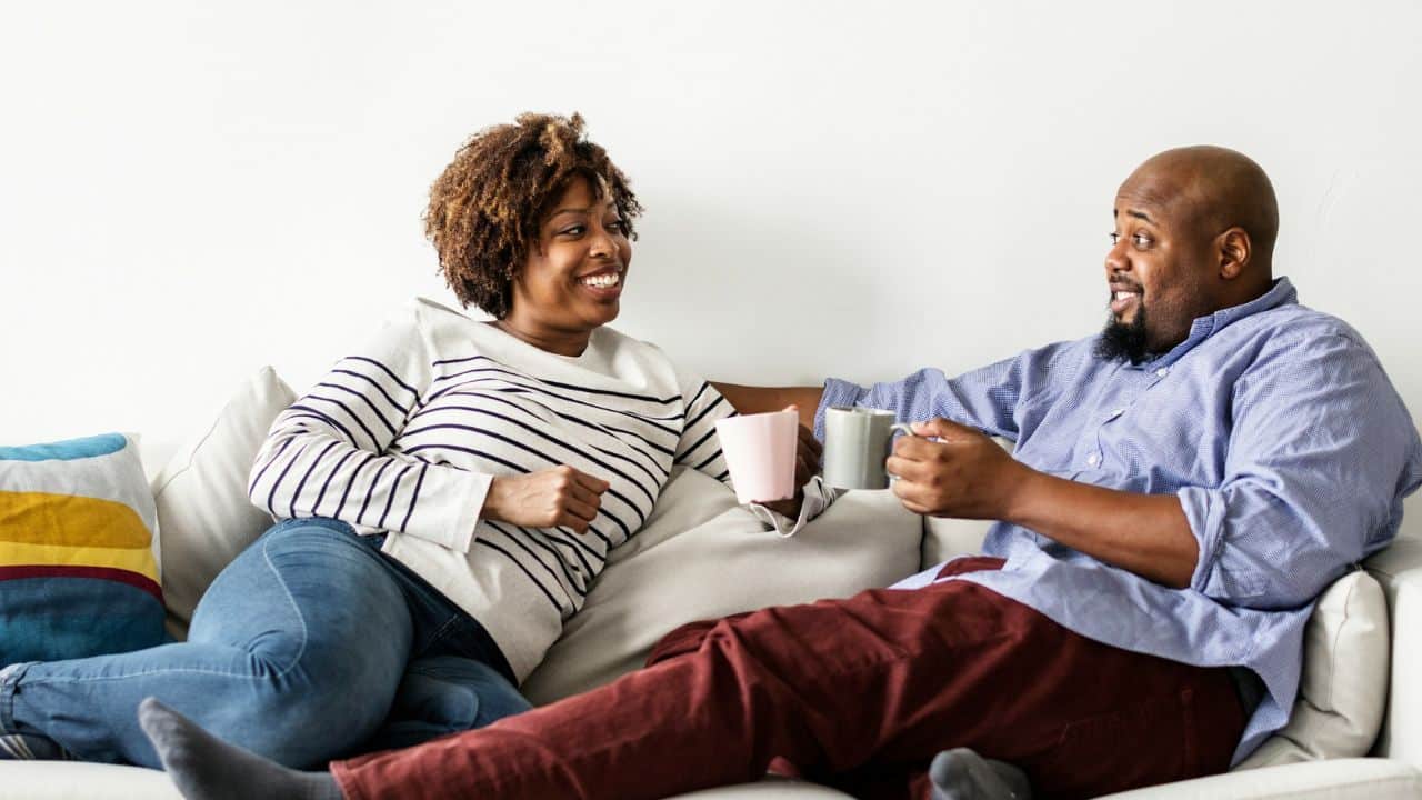 A happy Black couple sits on a couch, holding mugs and looking at each other.