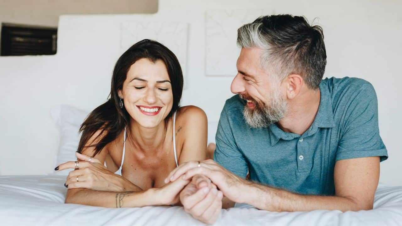 A happy couple lies on a bed, facing each other and holding hands.