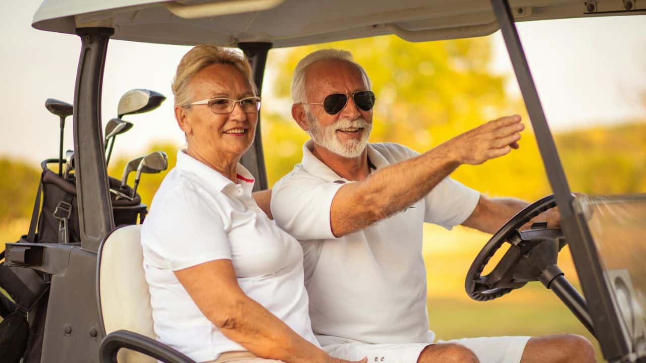 A senior couple smiles in a golf cart; the man points to something.