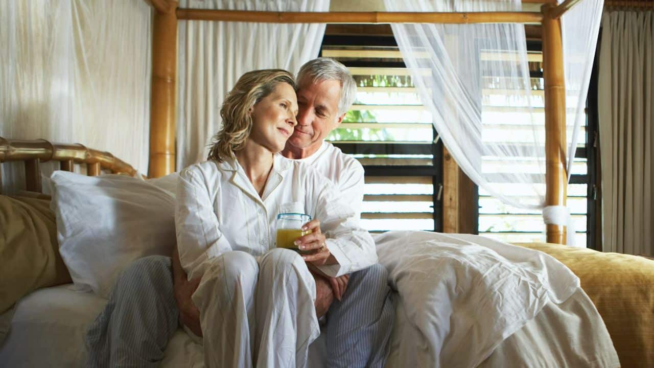 A happy man embraces a woman holding a drink in a canopy bed.