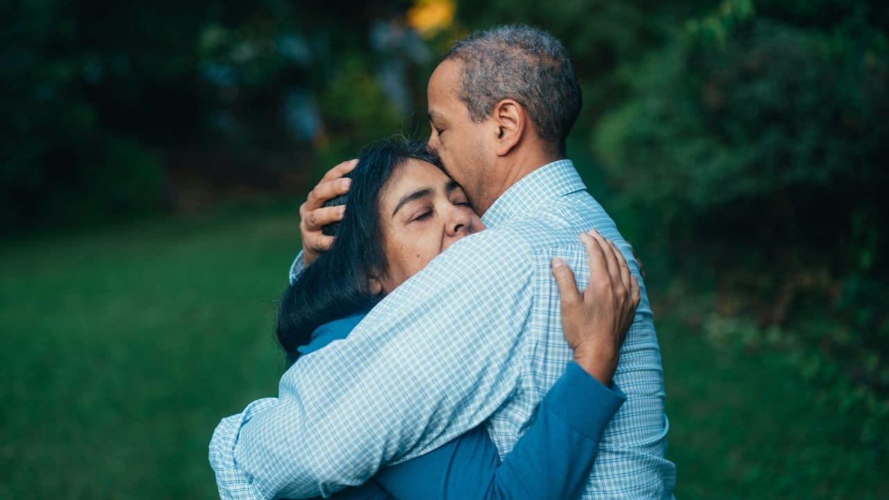 A man in a plaid shirt hugs a woman, kissing her forehead.