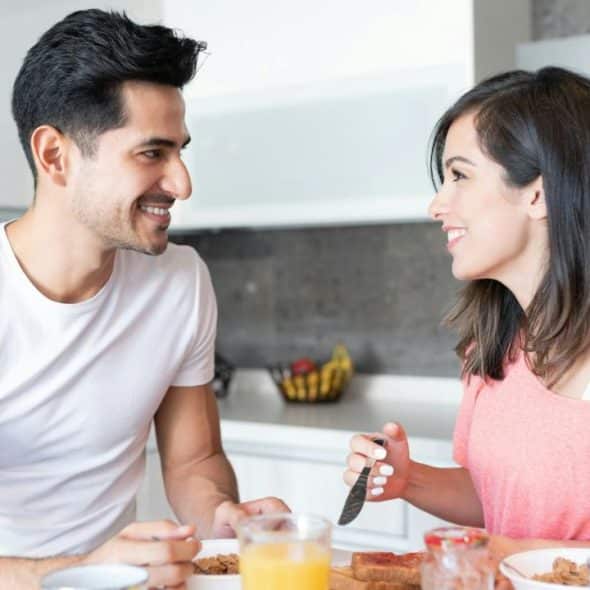 A man and a woman smile at each other while having breakfast at a kitchen counter.