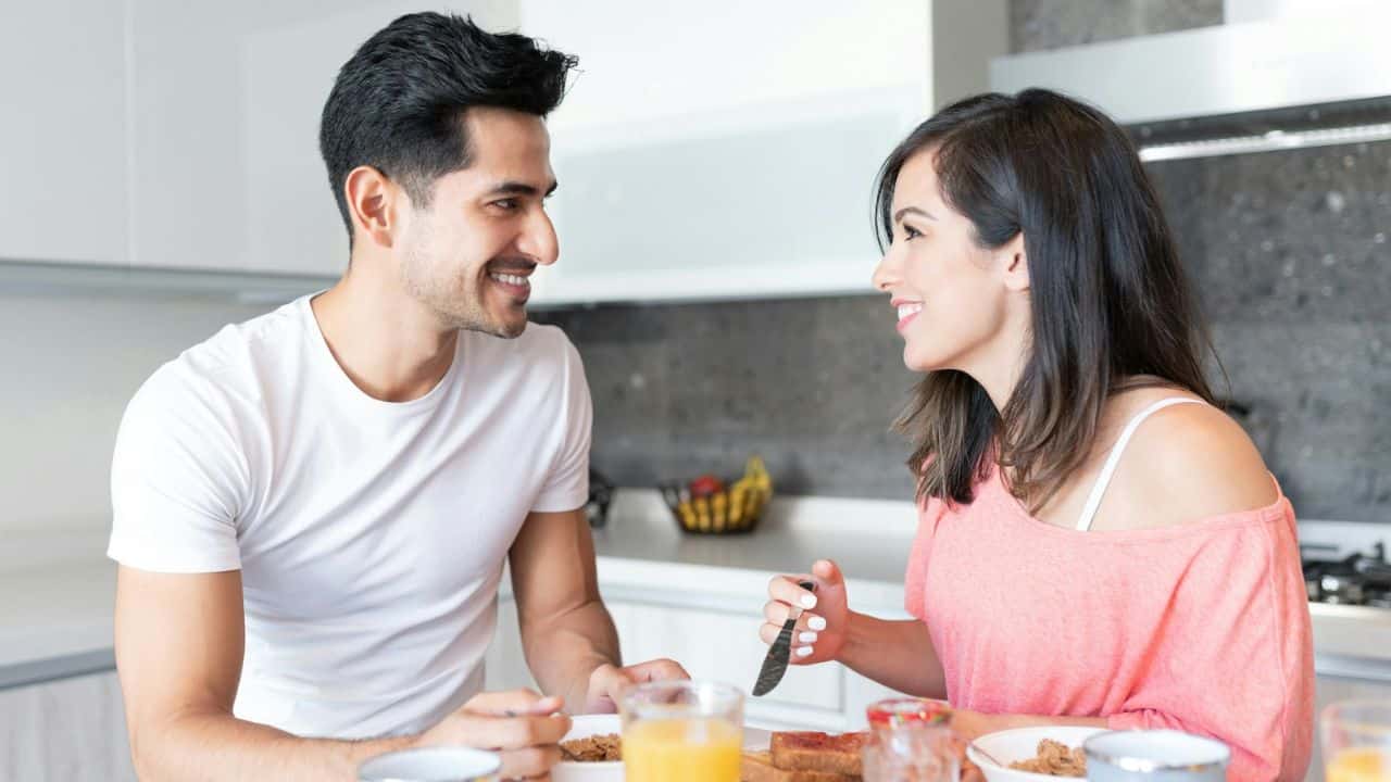 A man and a woman smile at each other while having breakfast at a kitchen counter.